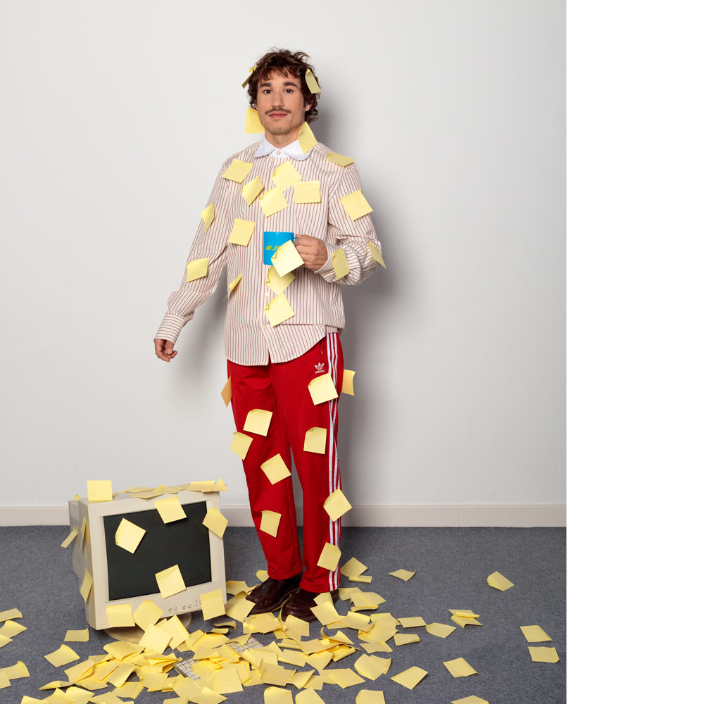 Plano general de Alberto Valero, viste una camisa de cuadros y un chandal rojo de Adidas. Está sujetando una taza de te, el cuerpo y el pelo está lleno de post-it amarillos. En el suelo hay una pantalla de ordenador antigua, de las de culo, también llena de post-it. En el resto de suelo de moqueta gris también hay más post-it