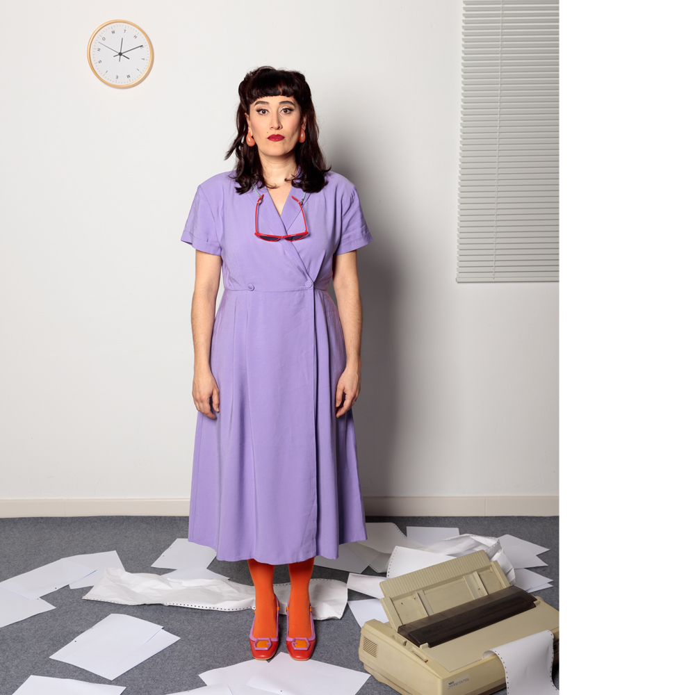 Plano general de susana Aguilera. Viste un vestido morado con medias rojas y zapatos a juego, le cuelgan unas gafas de sol también rojas. Sobre el suelo de moqueta gris el suelo hay papeles blancos desordenados y arrugados y una impresora, en la pared también hay un reloj y una persiana veneciana