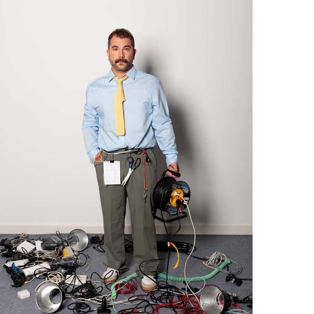 Plano general de Argider Aparicio. Viste una camisa azul, pantalón verde y corbata amarilla. Con la mano derecha metida en el bolsillo y la izquierda sujeta una alargador enrollable. A modo de cinturón un cable del que cuelgan tijeras, llaves, fiso y un blog de notas. El suelo de moqueta gris está lleno de cables y materiales electrónicos